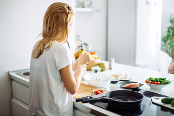 Rear view of blonde woman in white t-shirt preparing food for culinary blog. Young bloger female giving masterclass of cooking healthy food on kitchen