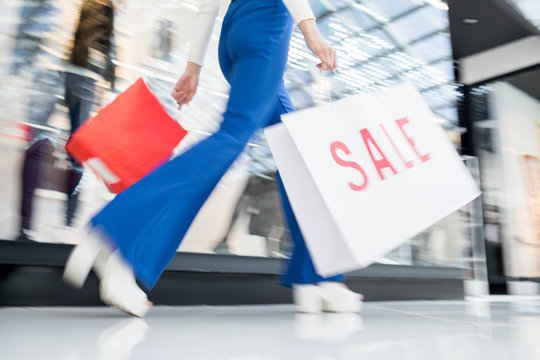 Unrecognizable woman in blue trousers waving shopping bags and walking over mall after visiting sale in stores