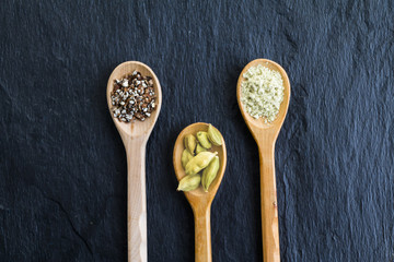 Different types of cardamom seeds - whole, high and low milling in wooden spoons on black slate background. Top view.