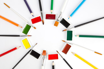 Solid watercolors pans with various paint brushes and colored pencils arranged in circle - top view with copy space on white background