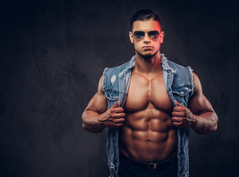 Sexy naked fashionable athletic man in a denim vest and sunglasses poses in a studio.