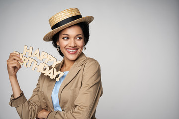 Beautiful smiling mixed race woman wearing canotier straw hat holding happy birthday text looking to side at blank copy space, isolated on gray background