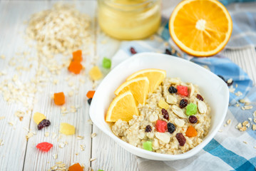 Homemade oatmeal porridge with honey, orange, raisin, peanut and candied fruit on white wooden background. Healthy breakfast.