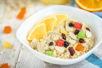 Homemade oatmeal porridge with honey, orange, raisin, peanut and candied fruit on white wooden background. Healthy breakfast.