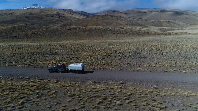 Aerial drone scene of van and trailer, motorhome in steppe, patagonia argentina riding on a gravel lonely road. Tromen volcano on the background. National Park. Camera moving left tracking car.