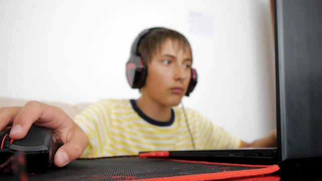 Teenager using laptop. Teen with headphones using wired mouse on pad. Close-up of hand moving computer gaming black and red mouse on mousepad