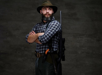 Portrait of a bearded hunter in a fleece shirt and hat standing with a rifle behind his back.