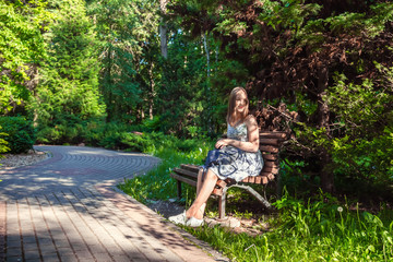 Girl in a dress and with a backpack sitting on a park bench in the summer day among the trees with copy space, the concept of lifestyle and recreation