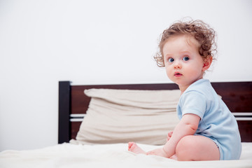 Curly little baby boy sitting on a bed in a bedroom.