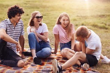 People, friendship and fun. Four men and women friends spend weekend outdoor, laugh at funny stories, have joyful expressions, sit on green grass, sunshine in background, enjoy summer vacation