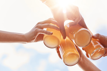 Summer, leisure, celebration and drinking concept. Close up shot of four hands clink bottles with lemonade or energy drink over blue sky and sunshine, celebrate starting vacations, have picnic - obrazy, fototapety, plakaty