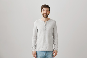 Smile attracts people. Portrait of good-looking pleasant bearded caucasian guy in loose pullover smiling broadly while standing over gray background with confident and friendly expression