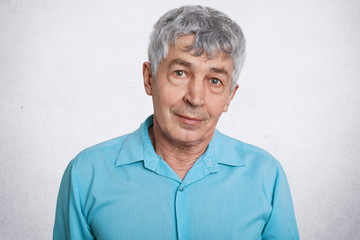 Portrait of attractive mature male with serious look, listens his grandson with great interest, dressed in shirt, poses against white concrete wall. Confidnet elderly man enjoys spare time at home