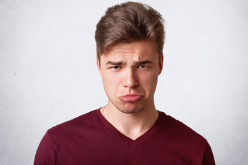 Fototapeta premium Photo of discontent gloomy male purses lower lip, looks with displeased expression, has sullen expression, going to cry after having big failure in life, expresses negative emotions, isolated on white