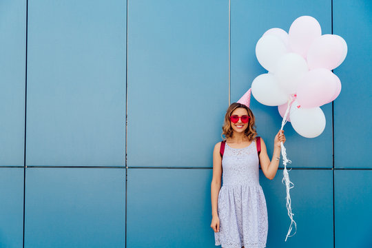 Cheerful Attractive Girl In Pink Sunglasses And Party Hat Holding Tender Air Balloons, Posing At Camera, Smiling. Dressed In Summer Dress. Outside.