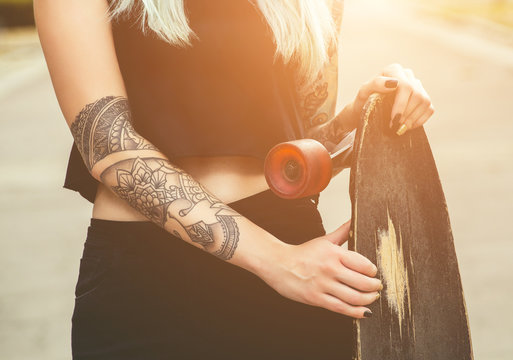 A beautiful, sexy hipster blonde with blue hair in tattoo stands with a longboard  on the street.