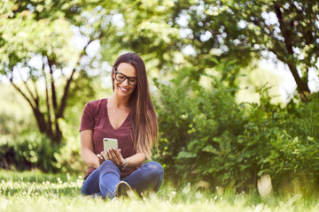 Fototapeta premium Smiling young woman sitting on ground in a park and using smartphone