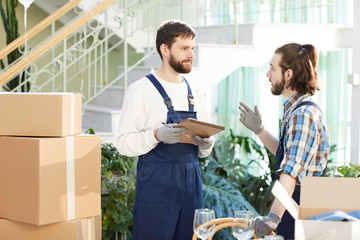 Busy confident handsome movers in overalls discussing work and thinking about next delivery order, serious young man with sketchpad attentively listening to colleague