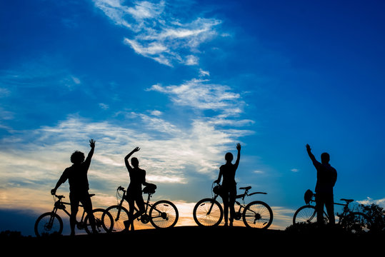 Silhouettes of young cyclists at sunset. Male and female persons with bikes waving with hands on evening sky background. Enjoying the infinity.