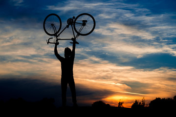 Silhouette of cyclist holding bike above head. Man lifting bicycle in the air on evening sky background. Beautiful evening scenery.