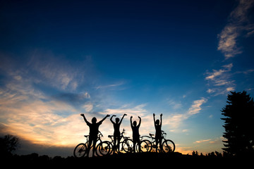 Fototapeta premium Friends with bikes at sunset background. Silhouettes of cyclists resting on evening sky background. Amazing summer evening scenery.