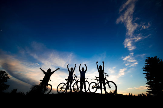 Cyclists with raised hands at sunset sky. Group of friends with bikes standing on hill at evening sky background. Enjoying of beauty.