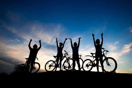Cyclists resting on hill at sunset sky. Group of young people standing with raised hands on hill on evening sky background. Carefree and freedom.
