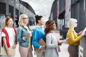 Group of multiethnic women in smart casual outfits standing in line while applying documents at registration for business event