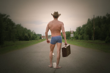 A naked man in shorts and a hat on his head and suitcase in his hands walks along an empty road....