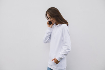 Smiling happy hipster girl wearing a white sweatshirt having a conversation via modern cellphone while standing on a white concrete wall background