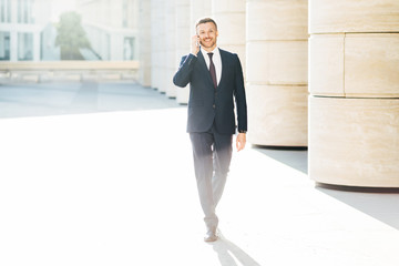 Horizontal shot of successful male entrepreneur in formal clothing, makes telephone call to partner via mobile phone, discuss creative ideas for common business, strolls outdoor over urban background