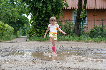 Naklejka premium Little girl is playing in the water in the middle of a ruined road, after rain