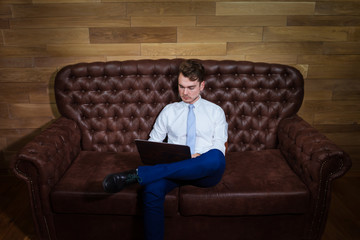 Serious man in suit is working with laptop at home office. Confident young manager sits on the leather sofa on background of wooden wall.