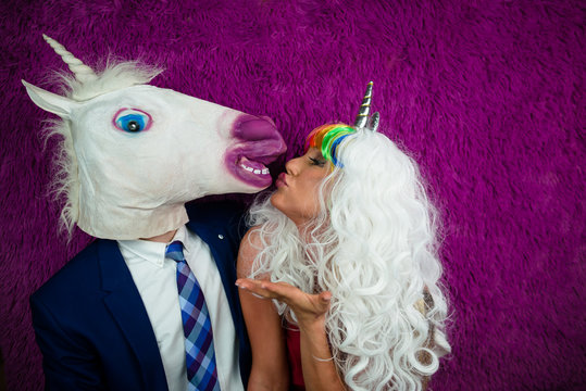 Portrait of unusual couple on the purple background. Freaky young woman in unusual wig and red dress is kissing funny man in comical mask and suit. Unicorn with girlfriend.