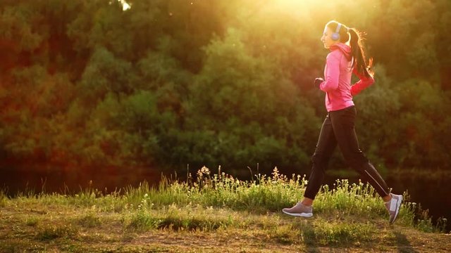 Brunette with long hair in headphones runs along the river in the Park in the morning at sunrise in the summer in a pink jacket and black pants