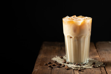 Ice coffee in a tall glass with cream poured over and coffee beans on a old rustic wooden table. Cold summer drink on a dark wooden background with copy space