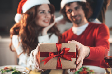 Happy New Year. Joyful young man and woman are holding gift together. They are sitting at table in kitchen and smiling. Focus on wrapped box with red ribbon 