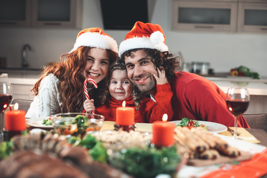 Christmas is our favorite holiday. Portrait of glad parents embracing their little child with love and tenderness. They are looking at camera and laughing while sitting at festive table 