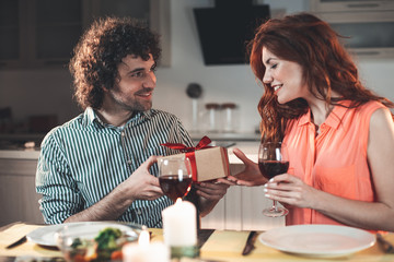This is for you. Happy man is giving present for his loving wife. Woman is looking at wrapped box with excitement and smiling. Lovers are sitting at table in kitchen 