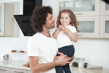 Caring father is spending time with his lovely daughter at home. He is holding a child and smiling while standing in cook room 