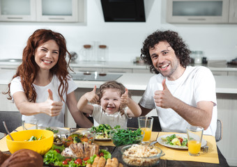 Everything is okay. Portrait of happy family is giving thumbs up while dining in kitchen. They are looking at camera and laughing 