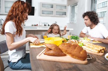 Friendly family is having breakfast together at home. They are sitting at table and smiling. Cute girl is holding fork and ready to eat salad 