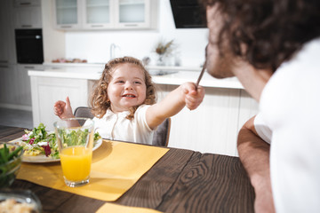 Taste this. Portrait of cheerful little kid is having fun with her dad in kitchen. Daughter is holding a fork and feeding a man with vegetable