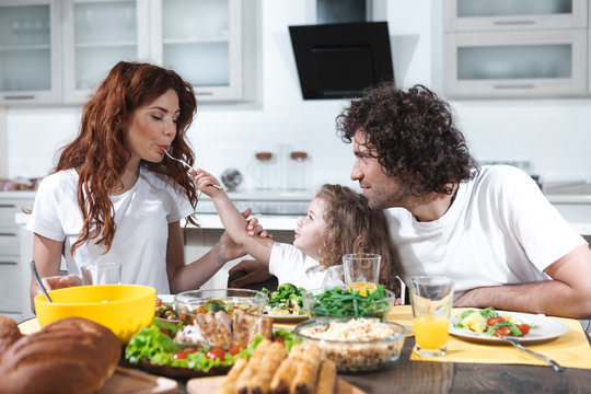 Cheerful little girl is feeding her mom by healthy food with joy. Woman is tasty dish with appetite. Man is looking at them with love and tenderness 