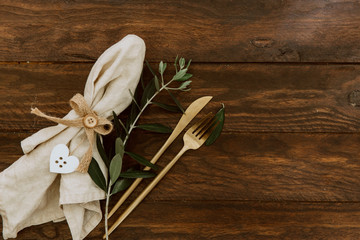 Gold cutlery with linen napkin and olive branch on wooden background. Wedding festive tableware