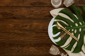 Gold cutlery with linen napkin and monstera leaf on wooden background. Wedding festive tableware