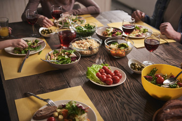 Close up of healthy dishes lying on wooden board. Family sitting together and eating vegetables, salads, rice, tomatoes, and drinking red wine