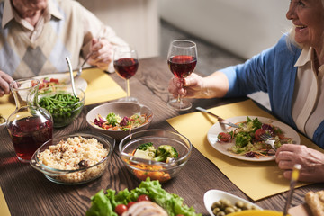 Close up of elderly woman hand holding red wine glass. She is sitting at table full of healthy dishes next to husband. Old female is raising goblet while eating salad with smile