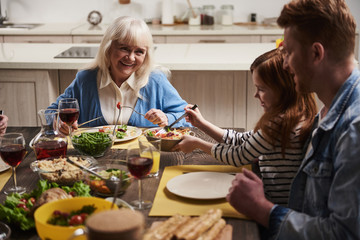 Three generations at one table. Happy grandmother talking to her dearest. Small girl is holding bowl with salad while her dad is sitting next to her. They are eating wholesome meal with joy