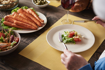 Close up of senior woman hands with cutlery. She is eating tasty nutritious food with ripe vegetables and wholegrain bread. Delicious meal is supplemented with a glass of red wine